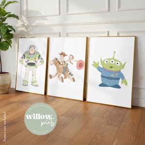 May include: Three framed art prints featuring characters from the Toy Story film. The prints include Buzz Lightyear, Woody and Jessie riding a horse, and the Little Green Alien. The frames have gold edges and are displayed on a wooden floor.