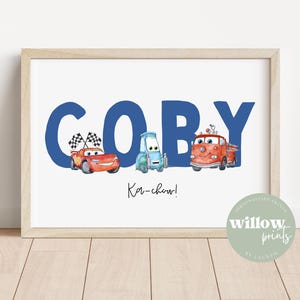 Può includere: Una stampa incorniciata con il nome "COBY" in blu, accompagnata da tre illustrazioni di auto dei cartoni animati. La frase "Ka-chow!" è scritta sotto. La stampa è in una cornice di legno chiaro.