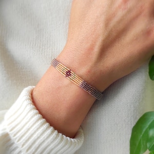 Puede incluir: Un brazalete de cuentas multicolores con una cuenta roja en el centro. El brazalete está en la muñeca de una persona.