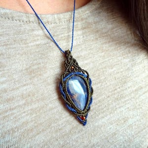 Könnte beinhalten: Eine handgefertigte Makramee-Halskette mit einem großen, tropfenförmigen blauen Edelsteinanhänger. Der Anhänger ist in eine gewebte Makramee-Fassung mit blauen und braunen Akzenten gefasst und hängt an einer zarten blauen Kordel. Ein einzigartiges Schmuckstück.