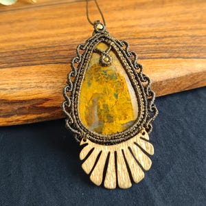 Puede incluir: Un collar de macramé marrón y dorado con un colgante de ágata amarillo y marrón. El colgante está rodeado por un marco de madera decorativo.