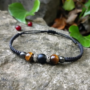 Peut inclure: Bracelet tressé noir orné de perles en œil de tigre et en onyx noir, avec des entretoises argentées. Le bracelet est monté sur un cordon noir et est présenté sur une surface en pierre grise. Bijou fait main.