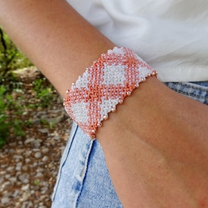 Op de afbeelding: Een kralenarmband met een geometrisch patroon van witte en koraalkleurige kralen. De armband wordt om de pols gedragen. De kralen zijn geregen met een metalen draad. Dit is een sieraad.