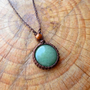 Puede incluir: Un collar con un colgante de piedra verde, un cordón marrón y cuentas de madera. El colgante está anudado con un diseño de macramé.