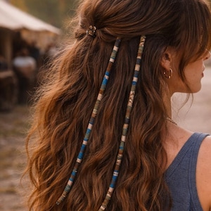 Puede incluir: Cabello castaño largo y ondulado peinado con una pequeña horquilla dorada y dos envolturas decorativas para el cabello. Las envolturas presentan patrones azules, verdes y marrones, agregando un toque bohemio al peinado.