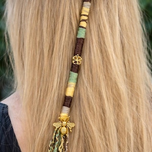 Puede incluir: Un accesorio para el cabello envuelto con un dije de abeja dorada y un dije de flor dorada. La envoltura para el cabello es marrón, amarilla, verde y beige. El accesorio está sujeto a cabello rubio largo.