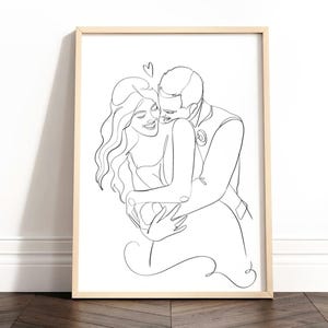 Op de afbeelding: Een ingelijste kunstdruk met een zwarte lijntekening op een witte achtergrond. De illustratie toont een koppel in een omhelzing in een doorlopende lijnstijl, waarbij de persoon links lacht en de persoon rechts voorover leunt, plus een klein hartje.