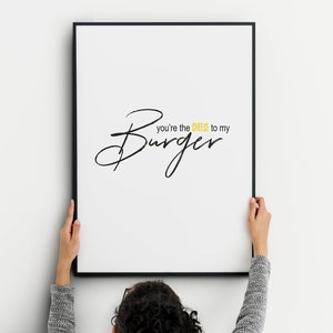 May include: A framed white print with black text that reads "you're the cheese to my Burger". The word "Burger" is in a large, cursive font. The word "cheese" is in a yellow font. The frame is black.