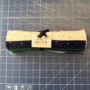 May include: A rolled bundle of fabric strips featuring various colors and patterns, including cream with small colorful stars, navy blue with white stars, and green with white dots. A paper label reads JOLLY BASICS COLLABORATIVE COLLECTION.