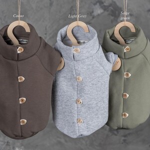 May include: Three pet sweaters in cacao, light gray, and sage green colors. Each sweater features a high collar, short sleeves, and a front placket with four light-colored wooden buttons. The sweaters are displayed hanging on wooden hangers.
