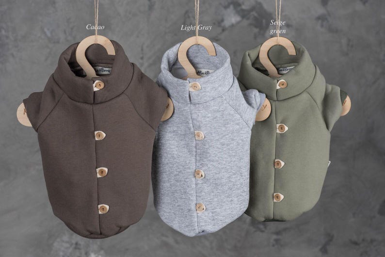 May include: Three pet sweaters in cacao, light gray, and sage green colors. Each sweater features a high collar, short sleeves, and a front placket with four light-colored wooden buttons. The sweaters are displayed hanging on wooden hangers.