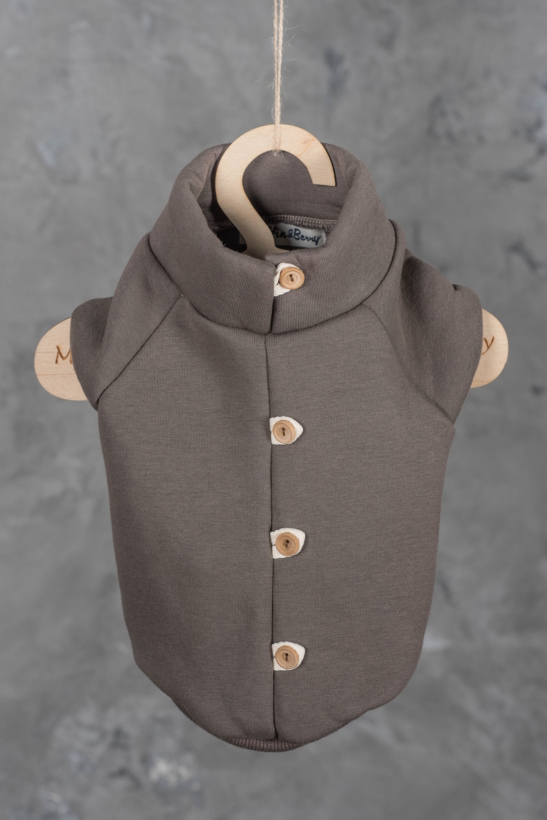 May include: A dark brown sleeveless vest with a high collar and four light brown wooden buttons down the front. The garment features white fabric accents behind each button and a small interior label that reads "Lin & Berry" at the neck.