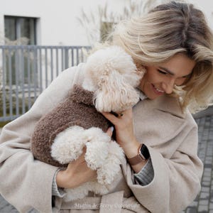 May include: A small, curly-haired dog wearing a brown, textured, sleeveless sweater. The dog is held by a person wearing a beige coat. The text Muffin & Berry copyright is visible at the bottom of the image.