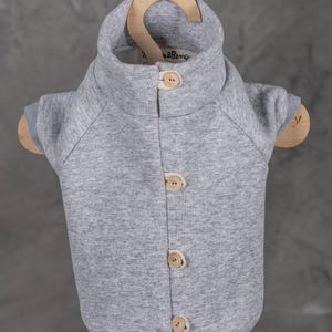 May include: A heather gray pet sweater with a high collar and four light-colored wooden buttons down the front. The garment features short sleeves with ribbed cuffs and a small interior label that reads "Twig & Berry" at the neckline.