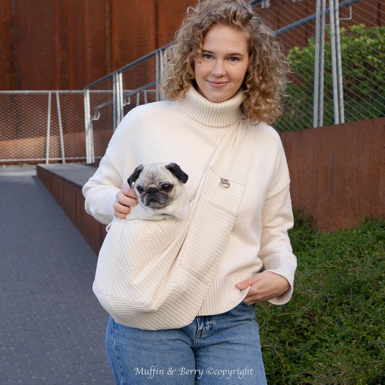 May include: A cream-coloured dog carrier with a waffle-weave texture, holding a pug with a black face and ears. The carrier is worn across the body. The dog carrier has a small metal logo on the strap.