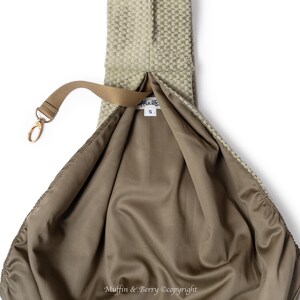 May include: A beige sling bag with a textured outer surface and a brown interior. The bag features a brown strap with a gold-coloured clasp. The interior lining is a smooth, satin-like material. The bag is open, showing the inside.