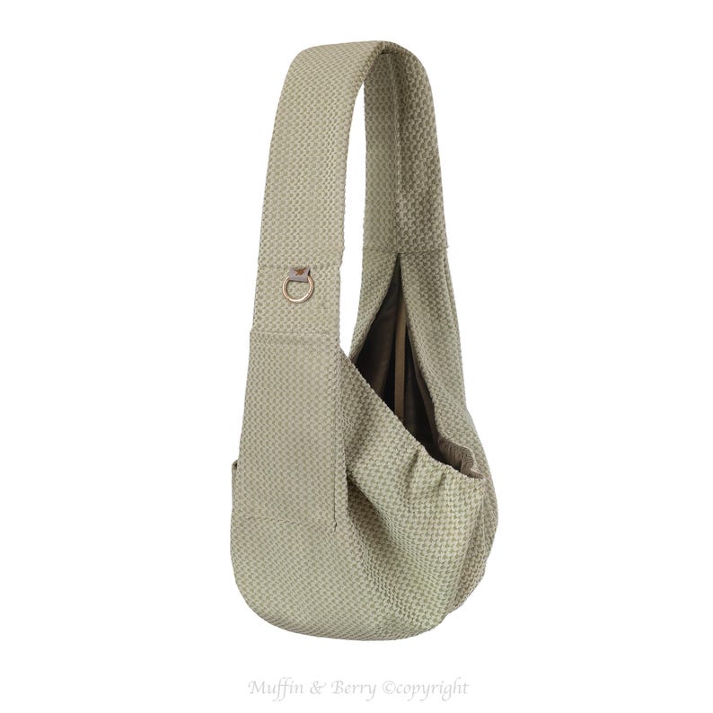 May include: A light green, textured sling bag with a single shoulder strap. The bag has a rounded shape with a deep pocket and a metal ring detail on the strap. The bag is designed to be worn over the shoulder.