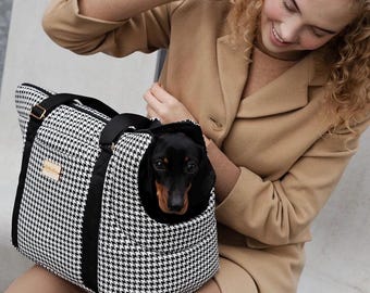 May include: A black and white houndstooth patterned pet carrier bag with black fabric handles and trim. The bag features a rectangular shape with a small tan rectangular brand label on the front and a black and tan dachshund dog inside.