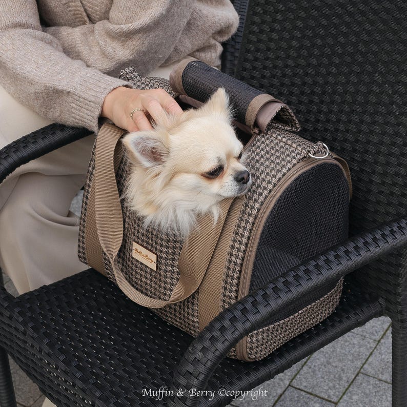 Puede incluir: Un transport&iacute;n para mascotas de color marr&oacute;n y beige con un peque&ntilde;o perro blanco en su interior. El transport&iacute;n tiene una ventana de malla y un asa. El perro est&aacute; mirando hacia afuera del transport&iacute;n.