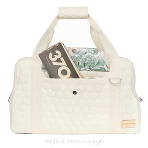 May include: A cream-colored quilted pet carrier with a hexagonal pattern, featuring two top handles, a front pocket containing a book with the number 370 on the cover, a floral patterned interior lining, and a small rectangular brand label on the bottom right.