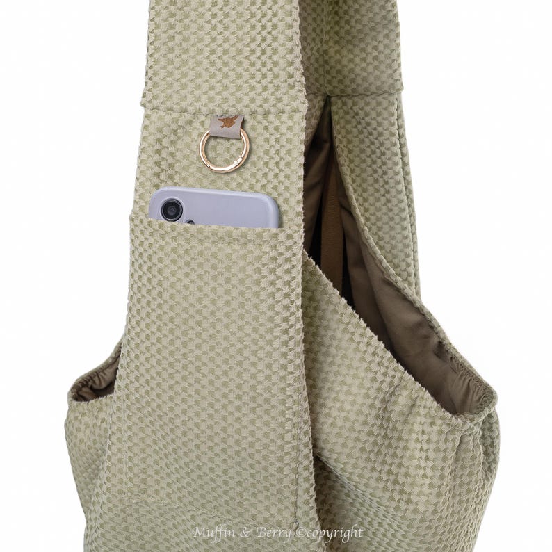 May include: A light green textured fabric sling bag with a pocket holding a smartphone. The bag has a gold-coloured ring and a small leather accent. The interior lining is a darker shade of green.