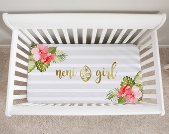 Op de afbeelding: Een witte wieg met een grijs en wit gestreept hoeslaken. Het laken heeft de tekst "neni girl" in gouden letters, een gouden embleem, roze en rode hibiscusbloemen en groene bladeren in de hoeken.