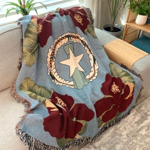 May include: A light blue woven throw blanket with a design featuring a white star, a wreath, and large maroon hibiscus flowers. The blanket has a fringed edge and is draped over a gray sofa.