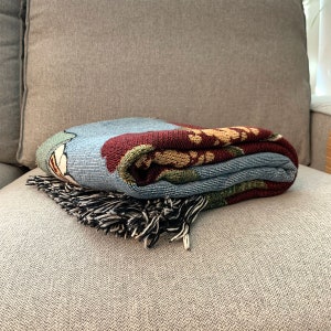 May include: Folded woven throw blanket with a floral design in shades of blue, red, green, and gold. The blanket has a fringe in black and white and is resting on a gray couch.