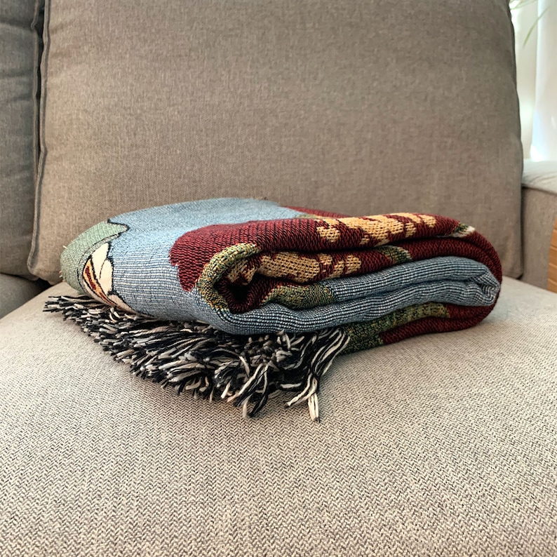 May include: Folded woven throw blanket with a floral design in shades of blue, red, green, and gold. The blanket has a fringe in black and white and is resting on a gray couch.