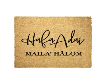 Op de afbeelding: Een bruine kokosvezel deurmat met de tekst "Hafa Adai" in een cursief lettertype en de tekst "Maila' Halom" in een sans-serif lettertype.