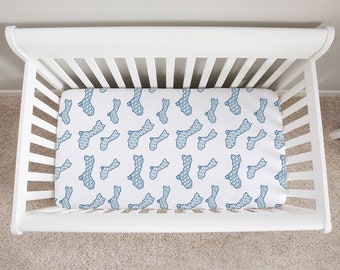 Op de afbeelding: Een witte wieg met een hoeslaken met een herhalend patroon van blauwe eilandvormen. De wieg is gemaakt van wit hout en staat op een lichtgekleurd tapijt. Het laken heeft een geometrisch ontwerp.