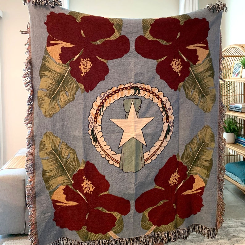May include: A light blue throw blanket featuring a design with four large maroon hibiscus flowers and green leaves. The center showcases a white star and a wreath design. The blanket has fringed edges.