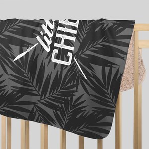 May include: A gray and black baby blanket with a palm leaf pattern. The blanket has the words "little chill" in white script with a white arrow graphic. The blanket is draped over a wooden crib.