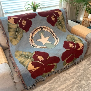 May include: A light blue throw blanket featuring large maroon hibiscus flowers and green leaves. The center design includes a white star, a wreath, and a ribbon. The blanket has a fringed edge and is draped over a gray sofa.