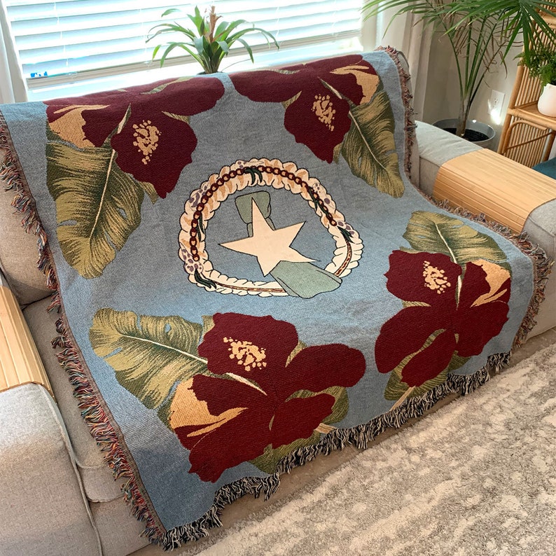 May include: A light blue throw blanket featuring large maroon hibiscus flowers and green leaves. The center design includes a white star, a wreath, and a ribbon. The blanket has a fringed edge and is draped over a gray sofa.