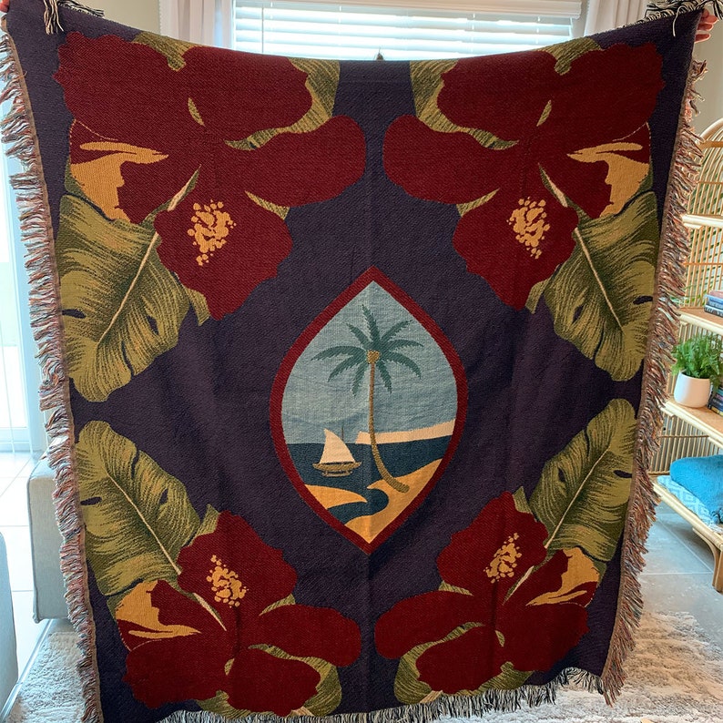 May include: A woven tapestry throw blanket with a navy blue background featuring a central design of the flag of Samoa. The flag is surrounded by four red hibiscus flowers with green leaves. The blanket has a fringed edge.