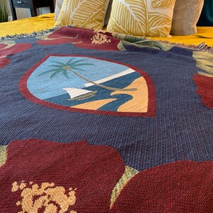 May include: A blue and red woven throw blanket with a large floral design and a small image of a palm tree on a beach in the center. The blanket has a fringed edge.