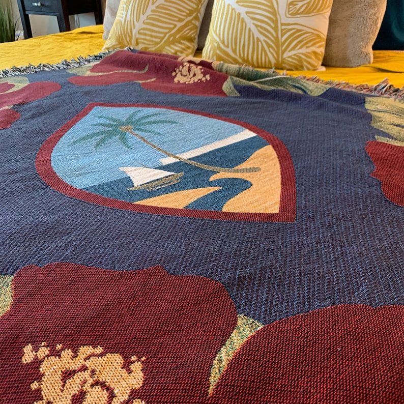 May include: A blue and red woven throw blanket with a large floral design and a small image of a palm tree on a beach in the center. The blanket has a fringed edge.