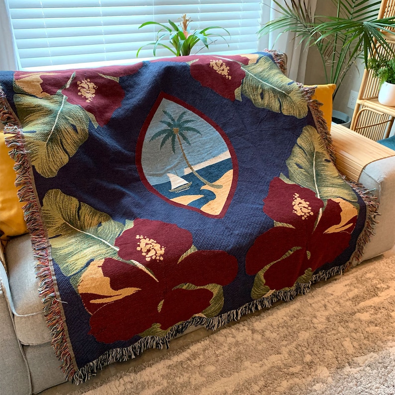May include: A woven throw blanket with a navy blue background featuring a red hibiscus flower design, green palm leaves, and a central crest with a palm tree and sailboat. The crest is surrounded by a yellow border.