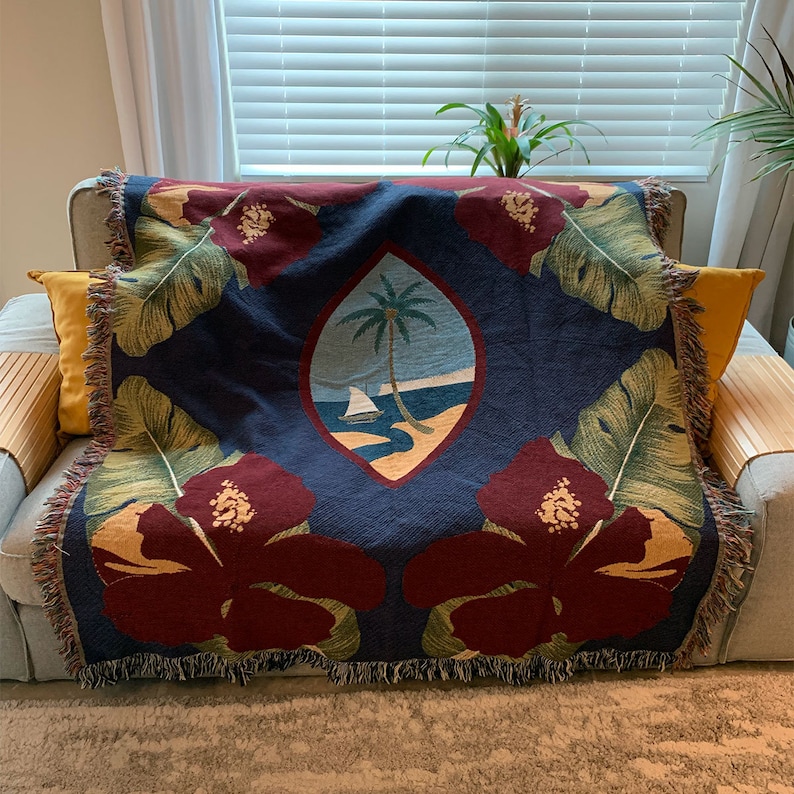 May include: A navy blue woven throw blanket with a red hibiscus flower design, green palm leaves, and a blue and white crest with a palm tree and sailboat. The crest is the national emblem of Samoa.