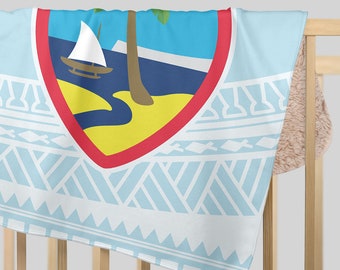 Op de afbeelding: Een lichtblauwe deken met het zegel van Guam, een rood omrand schild met een zeilboot, palmboom en eilandscène. De deken heeft een wit geometrisch patroon en is over een houten wieg gedrapeerd.