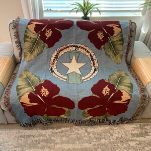 May include: A light blue throw blanket with a floral design featuring large maroon hibiscus flowers and green leaves. The center displays a white star within a wreath, with a light blue ribbon. The blanket has fringed edges.