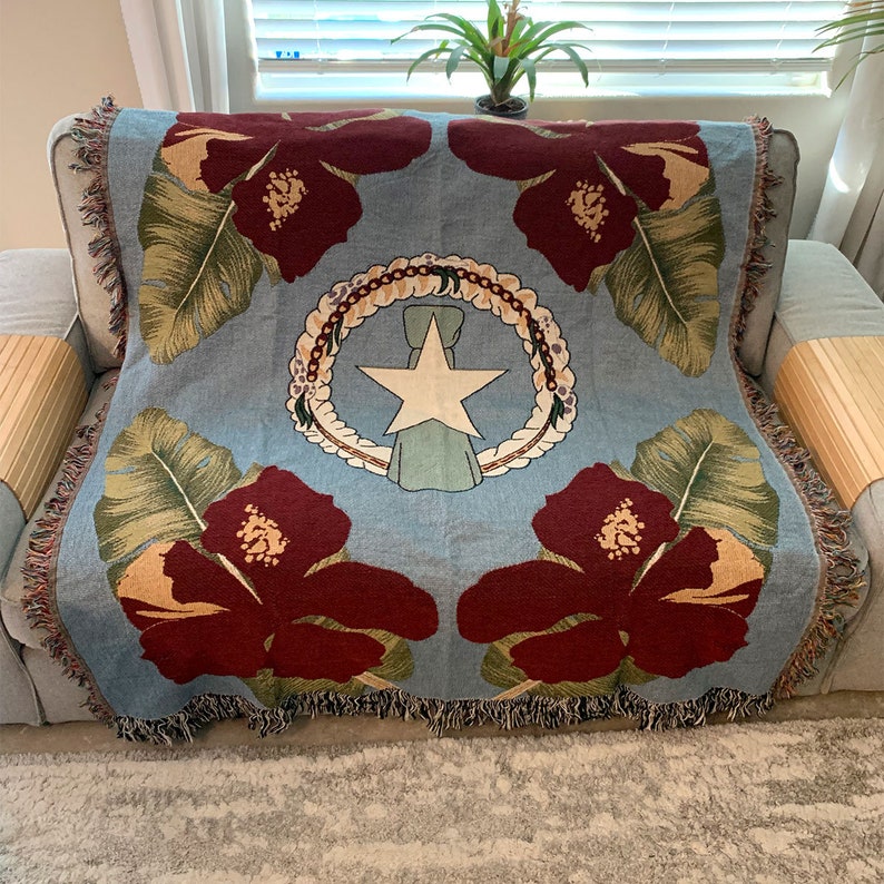 May include: A light blue throw blanket with a floral design featuring large maroon hibiscus flowers and green leaves. The center displays a white star within a wreath, with a light blue ribbon. The blanket has fringed edges.