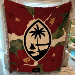 K&ouml;nnte beinhalten: Eine rote, gr&uuml;ne und wei&szlig;e gewebte Wolldecke mit einem schwarzen Wappen, das einen Palmenbaum und ein Segelboot zeigt. Das Wappen ist von Hibiskusbl&uuml;ten und gr&uuml;nen Bl&auml;ttern umgeben.