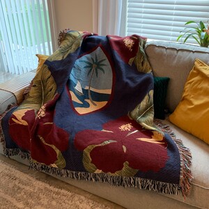 May include: A navy blue throw blanket with a woven design featuring palm trees, hibiscus flowers, and a sailboat. The blanket has a fringe border and is draped over a couch.