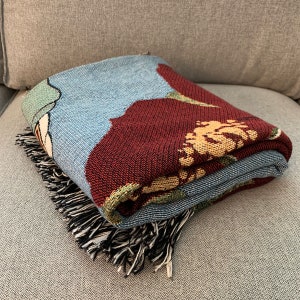 May include: Folded woven throw blanket with a floral design in shades of blue, burgundy, and beige. The blanket has a black and white fringe along one edge. The blanket is folded on a gray surface.