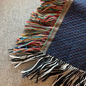 May include: Close-up of a blue and black woven throw blanket with colorful fringe. The fringe is made of multiple colors of yarn, including red, yellow, green, and blue.