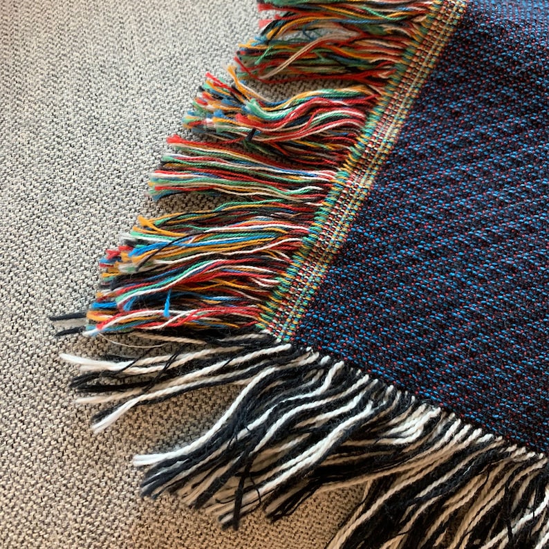 May include: Close-up of a blue and black woven throw blanket with colorful fringe. The fringe is made of multiple colors of yarn, including red, yellow, green, and blue.
