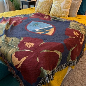 May include: A woven throw blanket with a navy blue background and a large red hibiscus flower design. The blanket features a central crest with a palm tree and a sailboat on a beach.