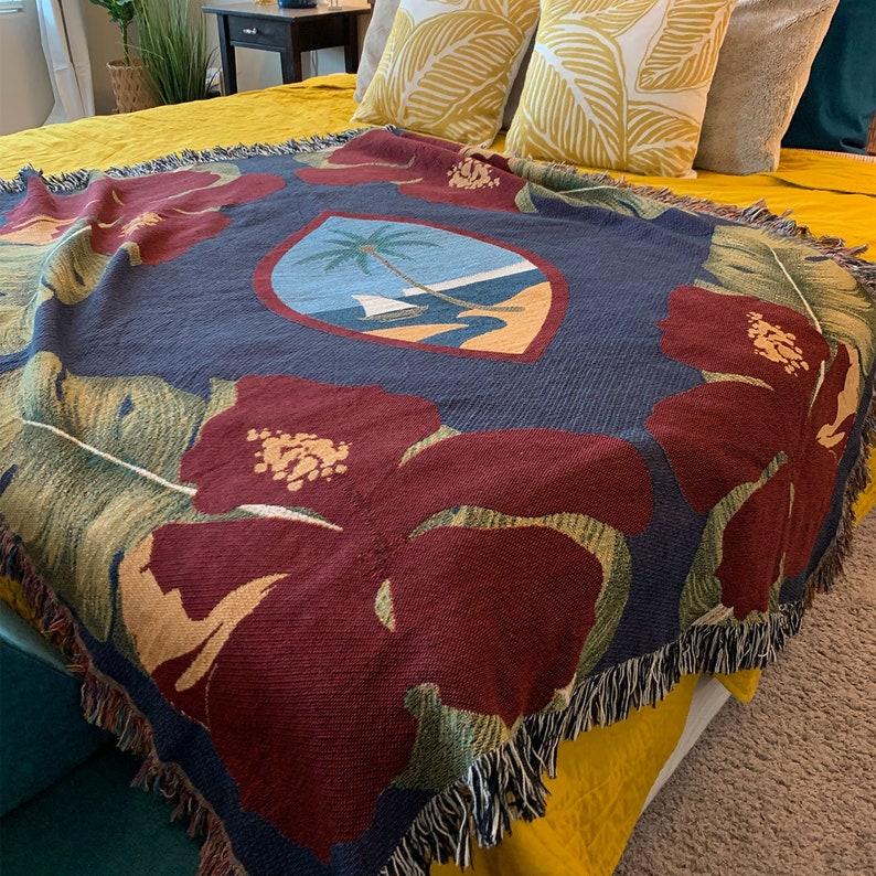 May include: A woven throw blanket with a navy blue background and a large red hibiscus flower design. The blanket features a central crest with a palm tree and a sailboat on a beach.
